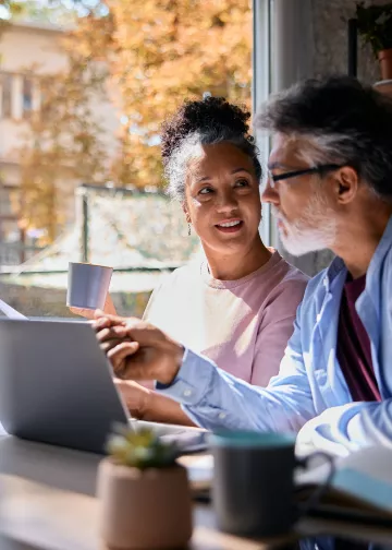  Two people collaborating on a laptop at a table near a window, one holding a coffee mug, engaged in warm conversation
