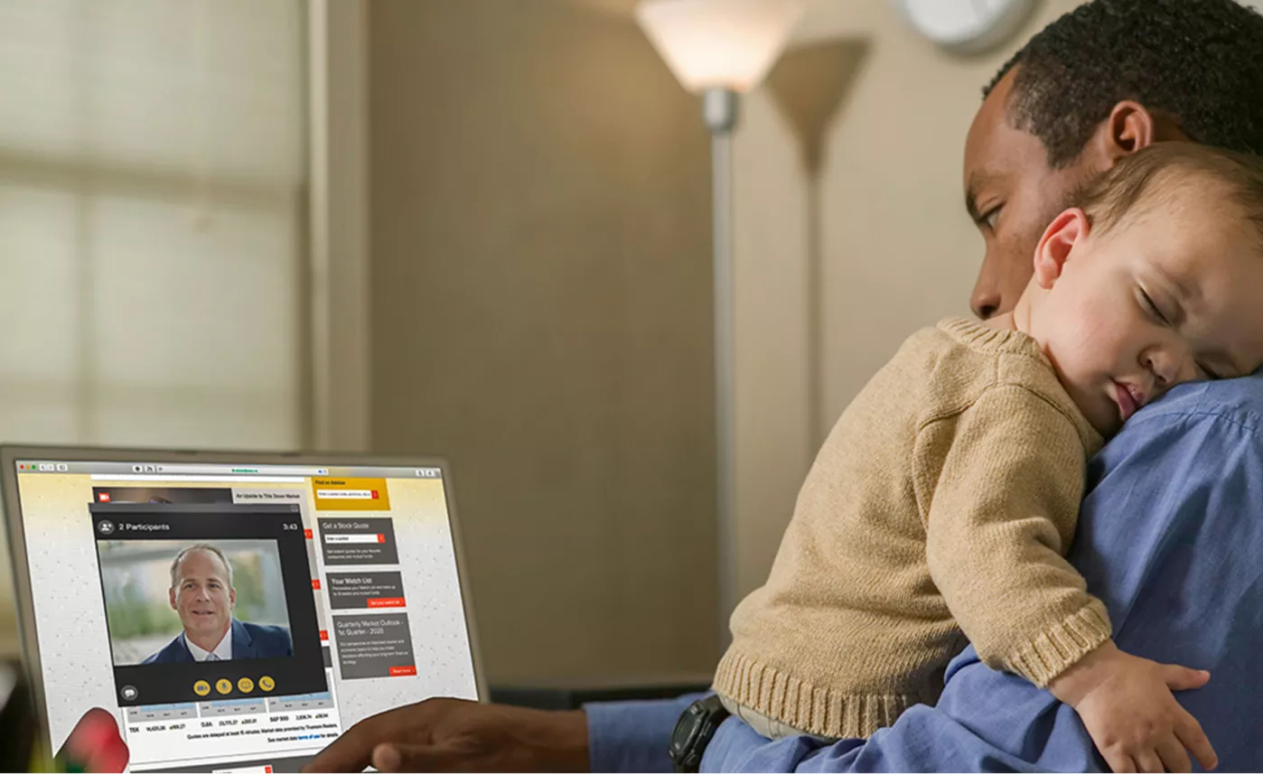  A young father talks to an Edward Jones advisor on a web conference in his home while his infant son sleeps on his shoulder.
