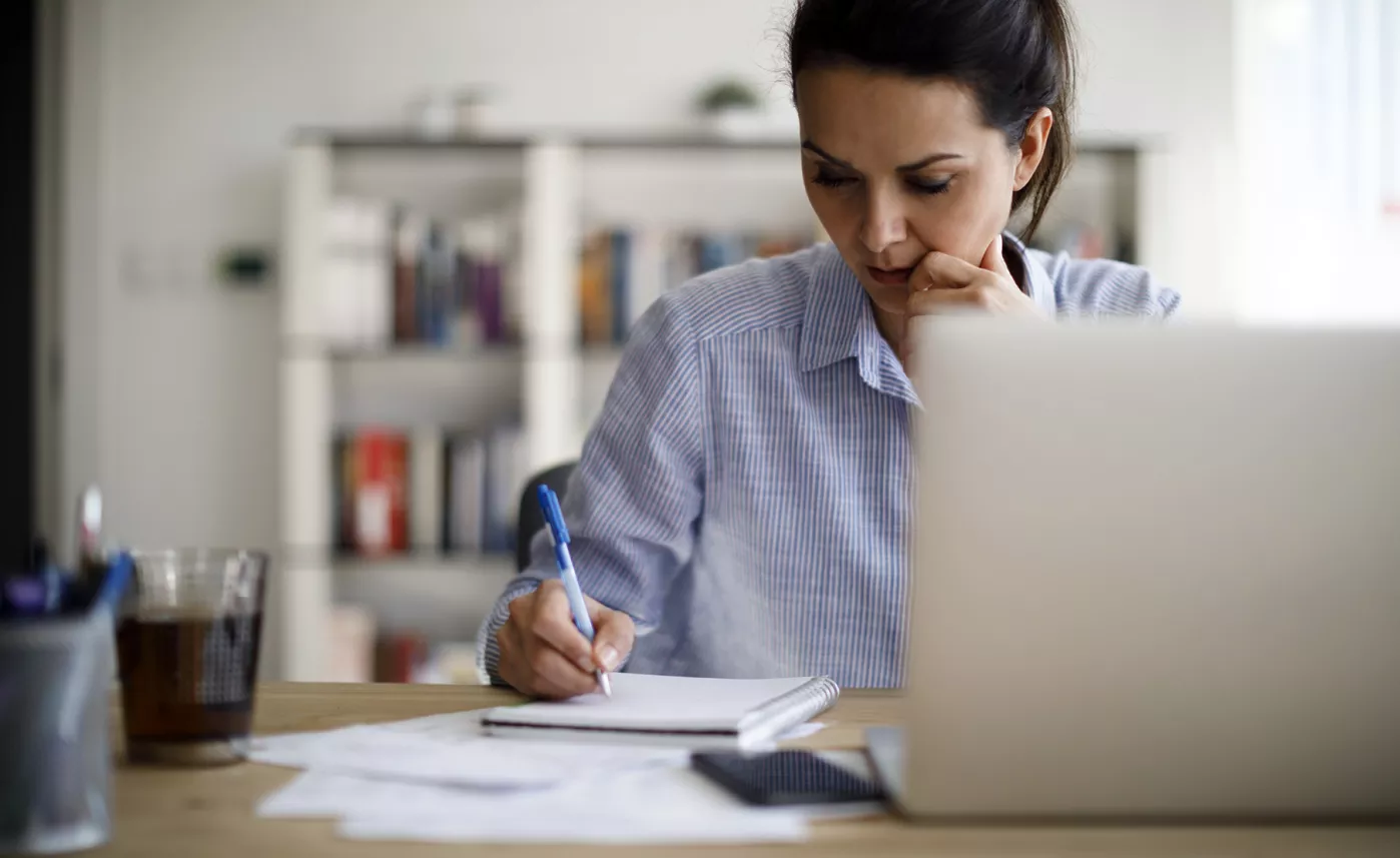  Une femme assise devant son ordinateur portable écrit dans un carnet et calcule soigneusement sa stratégie financière.
