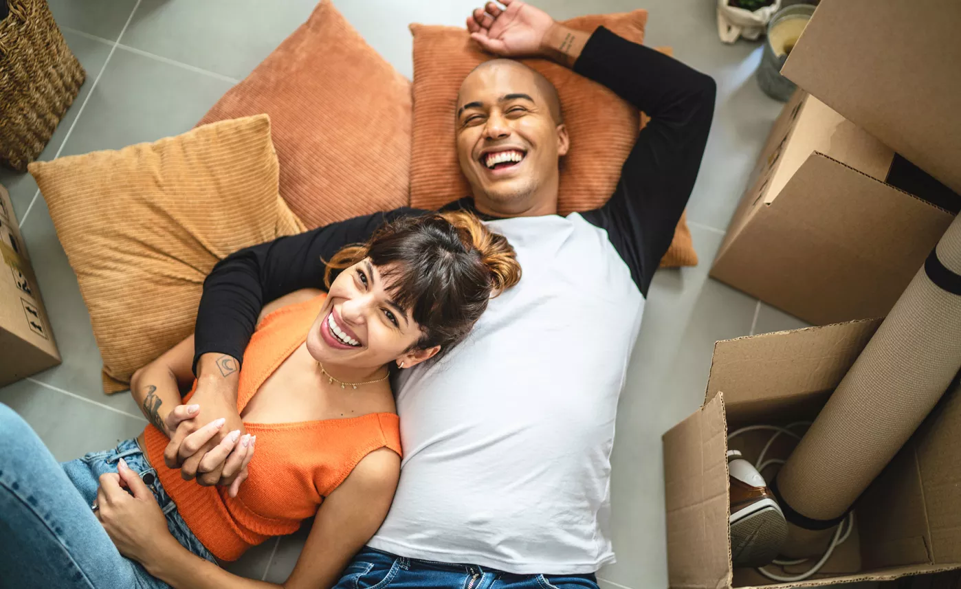  Aerial shot of a couple laying on the floor on pillows, laughing, surrounded by open moving boxes.
