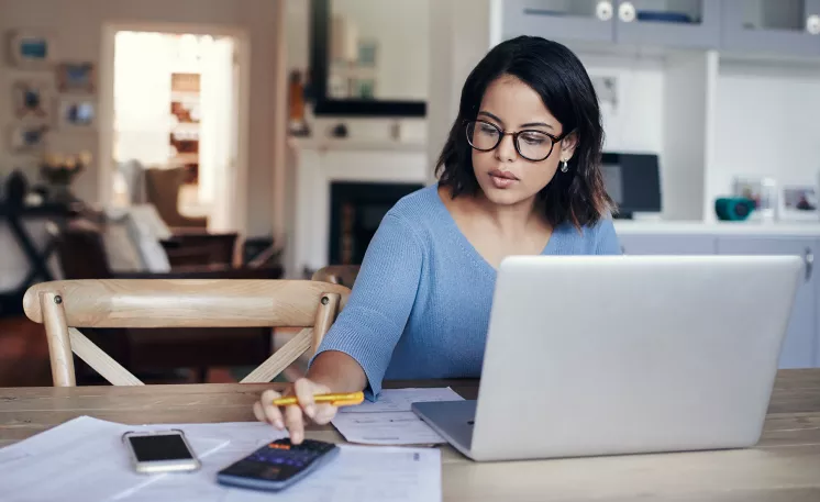 A person sitting at a kitchen table in front of their laptop, typing on their phone.
