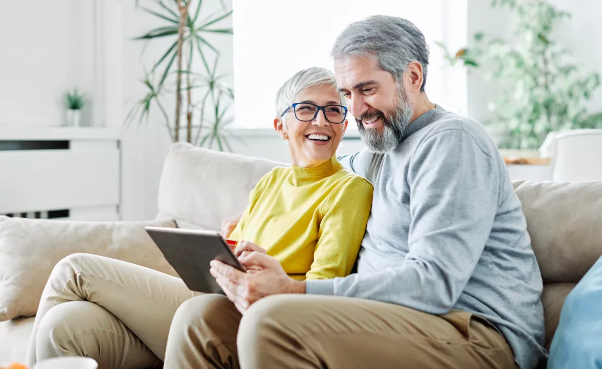  Un couple à l’âge de la retraite sourit en lisant sur une tablette sur le divan.

