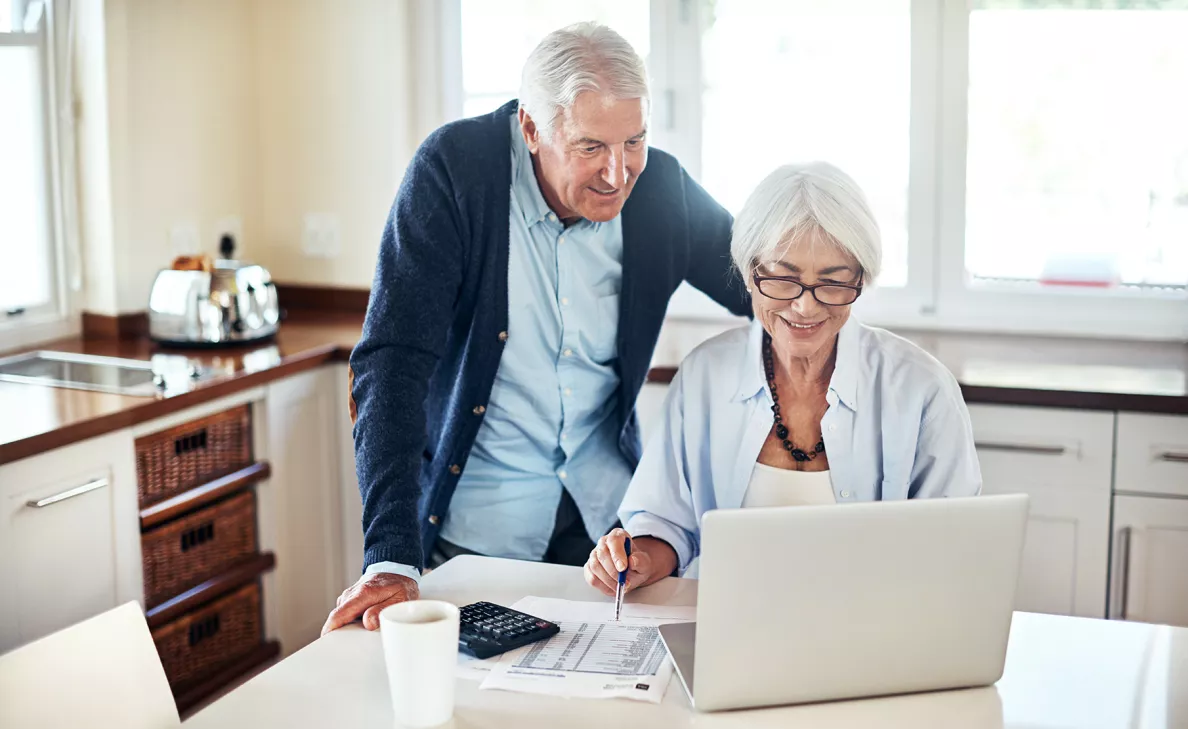  A retirement-aged couple check their account on a laptop at their kitchen table.
