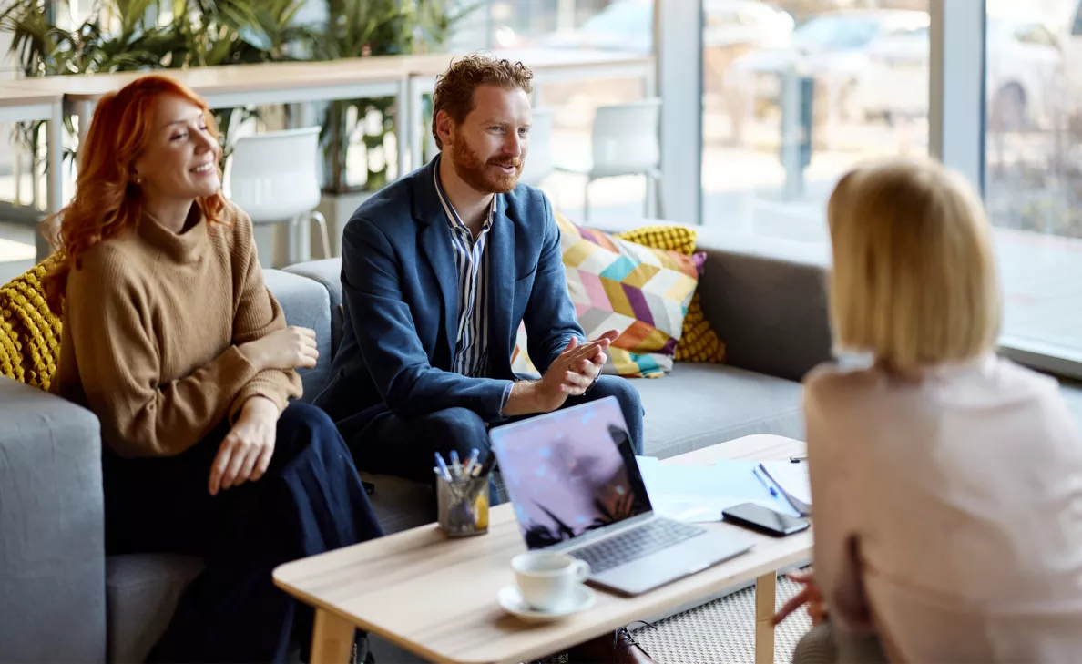 Two people, sitting across from a third person while chatting over a cup of coffee and a laptop.
