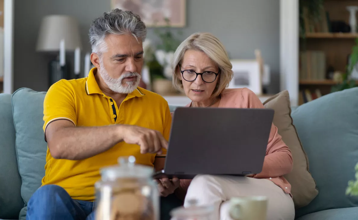  couple sitting closely together on a couch, both looking at a laptop screen with interest. The room has a cozy atmosphere, suggesting they are at home, possibly browsing or working together.
