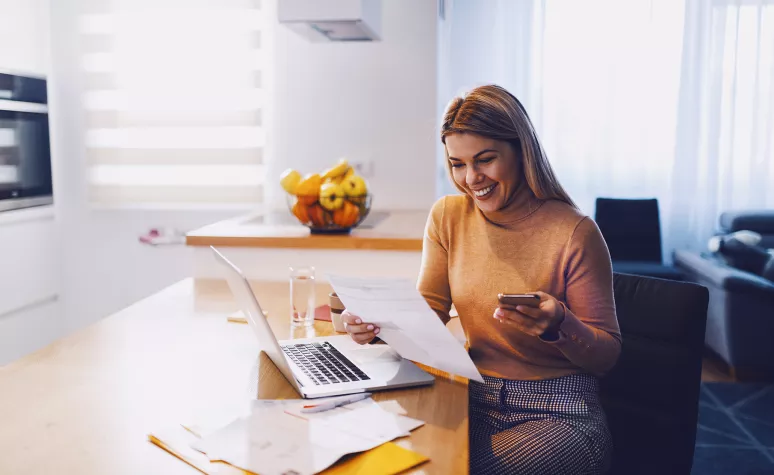 Une femme assise devant son ordinateur portable ouvert sourit en lisant des documents imprimés.