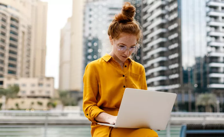 Une jeune femme travaille sur son ordinateur portable tout en étant assise à l’extérieur dans une zone urbaine.