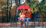 De jeunes parents s’assoient sur un banc de parc avec leur petite fille sous un parapluie alors qu’il pleut.