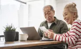 A retirement-aged couple review financial documents with their laptop open on their kitchen table.