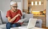 An elderly woman reviews printed documents in front of her laptop.