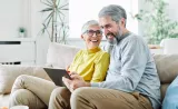 A retirement-aged couple smiles as they read from a tablet on the couch.