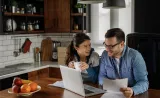 A young couple reviews retirement documents and their retirement account on their laptop in their kitchen.