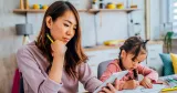 Parent and young child sitting at table. The parent is looking at their phone in contemplation while the child colours.