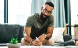 A man sitting on his couch writing in a notebook on his coffee table
