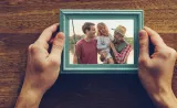Hands holding a framed picture of laughing parents with a child in front of a lake. One parent holds a fishing rod, while the other parent holds the child.