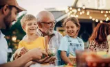 Four people outdoors at an evening event. One person on the left is serving food from a bowl. In the center, a grandparent wearing glasses is smiling while holding two children.
