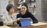 A young woman sits with her grandmother as they look at financial information on a tablet together.