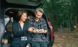 Deux personnes assises dans le coffre ouvert d'un VUS en forêt, riant et regardant un téléphone intelligent ensemble lors d'une aventure en plein air.