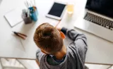 A young child works on their homework at a desk with a laptop open in front of them.